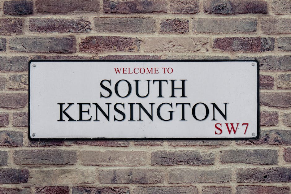 A rectangular white street sign mounted on a brick wall displays the text 'Welcome to South Kensington' in black lettering, with 'SW7' in red in the bottom right corner. The brick wall behind the sign features reddish-brown bricks with visible mortar joints, providing a textured background. The sign is situated outdoors, likely attached to the exterior of a building, and is evenly lit, highlighting its clear, legible text. The natural setting and urban environment are consistent with a residential or commercial area in South Kensington, London. This visual element is relevant to house removals or relocation services by Man with Van South Kensington, as it reflects the location context for home or furniture moving activities within this district, emphasizing the importance of local knowledge and logistics in moving operations.
