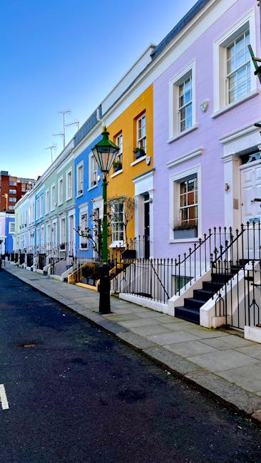 A wide street scene in South Kensington featuring a row of elegant Victorian-style terraced houses with cream-colored facades, decorative cornices, and tall windows. The buildings are several stories high and have small balconies with black railings, with some featuring flags hanging from the entrances. The street is busy with pedestrians walking on the pavements, including a man and woman, and a cyclist riding along the roadway. Parked cars are present along the curb, and a construction crane can be seen in the background, indicating ongoing development. The sky is partly cloudy with patches of blue, and the overall setting suggests a typical day of home relocation or furniture transport, with [COMPANY_NAME] providing removals services in the area, supporting efficient packing and moving processes. Visible elements such as the street, parked vehicles, and the surrounding architecture align with typical house removals in South Kensington.