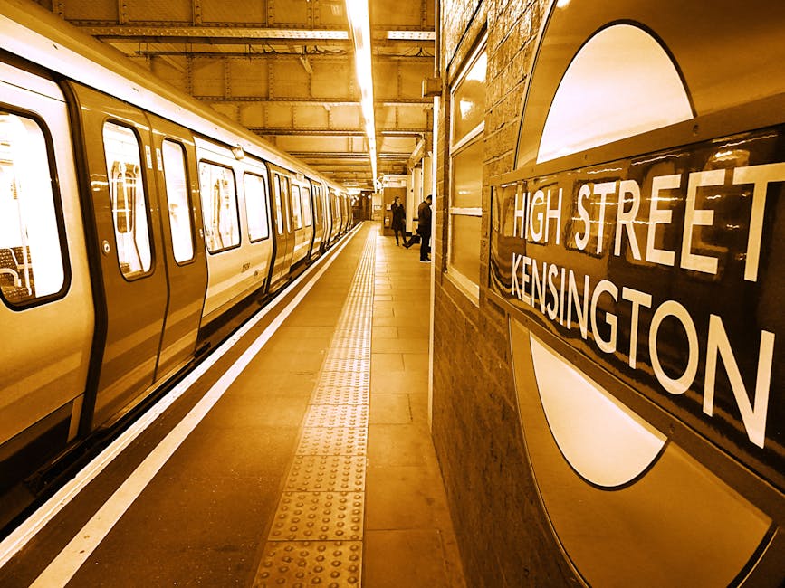 Inside a London underground station at High Street Kensington, the platform features a yellow train with multiple windows, some showing reflections of the interior. The platform surface is tiled, with tactile paving strips near the edge for accessibility. Two passengers are visible in the distance, standing near the platform edge, possibly waiting for the train. The station wall on the right displays a round sign with a black border and a white background, bearing the station name 'High Street Kensington' in bold black letters. Overhead lighting illuminates the space with a warm, yellowish glow. This scene captures the typical environment for house removals or furniture transport logistics involving safe loading and unloading of items during a home relocation process, with proximity to station facilities supporting efficient travel or transport arrangements. Man with Van South Kensington uses such settings for assisting clients with relocation and moving services in the area.