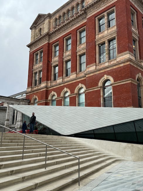 A modern, curved glass entrance extension attached to a historic red-brick building with classical architectural details, situated at the top of an outdoor marble staircase with metal handrails. Two individuals are standing on the staircase near a red carpeted area, likely preparing for a home relocation or furniture transport process. The scene shows an overcast sky, with the building's windows and stone detailing visible, reflecting a mix of historical and contemporary design typical of prominent properties in Museum district, South Kensington. This setting relates to the logistical aspects of house removals, where careful loading and unloading of furniture and packing materials take place during a move coordinated by Man with Van South Kensington, experts in removals near Exhibition Road.
