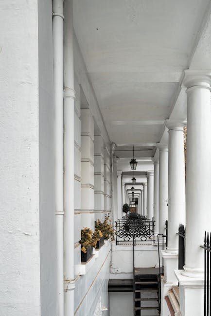 A view of a narrow outdoor corridor on Gloucester Road in South Kensington, featuring white-painted columns and walls supporting the building's façade. Several black iron railings line the edge of the balcony, with potted plants placed along the ledge. The corridor is illuminated by hanging lantern-style light fixtures, and the area appears clean and well-maintained, with a staircase descending to the lower level. This setting illustrates the typical architecture of a residential property undergoing home relocation, where furniture and boxes might be transported through stairs and lift access during a comprehensive moving process. The image reflects the environment handled by property removals specialists like Man with Van South Kensington during packing, loading, and furniture transport activities as part of house moving services.