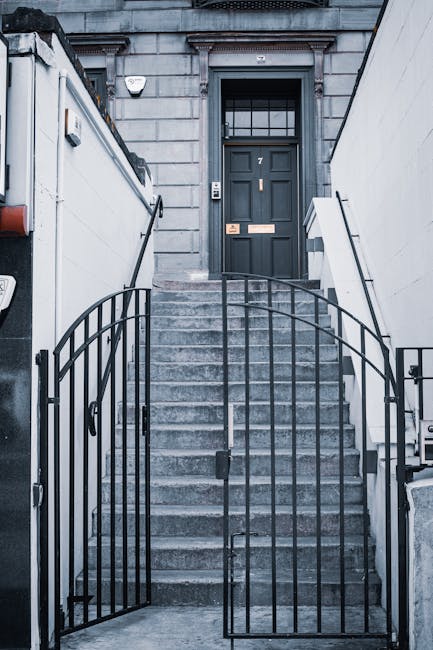 A view of a narrow outdoor corridor on Gloucester Road in South Kensington, featuring white-painted columns and walls supporting the building's façade. Several black iron railings line the edge of the balcony, with potted plants placed along the ledge. The corridor is illuminated by hanging lantern-style light fixtures, and the area appears clean and well-maintained, with a staircase descending to the lower level. This setting illustrates the typical architecture of a residential property undergoing home relocation, where furniture and boxes might be transported through stairs and lift access during a comprehensive moving process. The image reflects the environment handled by property removals specialists like Man with Van South Kensington during packing, loading, and furniture transport activities as part of house moving services.