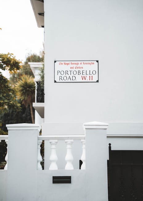A white exterior wall of a residential property features a sign indicating it is located on Portobello Road within the Royal Borough of Kensington and Chelsea, with a red and black address number W.11. In front of the wall, there is a small white fence with classical-style balustrades, partially obscuring a black mailbox or utility box. To the left, a portion of a balcony with a black wrought iron railing is visible, along with leafy green trees and shrubs providing a natural backdrop. The scene is well-lit with natural daylight, highlighting the clean lines and smooth finish of the building's exterior. This setting is typical for house removals or relocation services, where careful handling of furniture or boxes might occur near the property entrance or on the pavement outside, supporting the context of moving logistics organized by companies like Man with Van South Kensington, especially when complying with local council loading bay rules for removals.