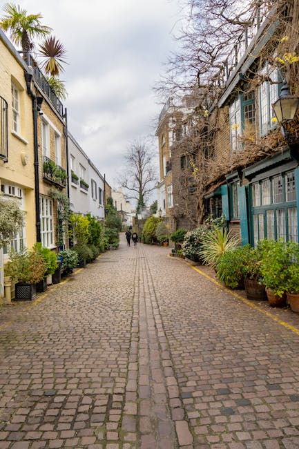 A cobbled street of a residential area in South Kensington during daytime, lined with multi-storey houses on both sides. The houses feature a variety of architectural styles with large windows and decorative elements; some with balconies, others with glass extensions or verandas. Potted plants and small trees are placed along the pavement outside each property, adding greenery to the scene. The sky overhead is partly cloudy, with bare trees indicating a possible early spring or late autumn. In the distance, two people are walking along the street, while a man from Man with Van South Kensington is seen loading or unloading furniture and boxes onto or from a large van parked on the right side. This scene captures a typical home relocation process, including the loading of packing materials like cardboard boxes, wrapped furniture, and protective padding. The environment suggests an organised, professional moving operation involved in furniture transport and packing and moving services.