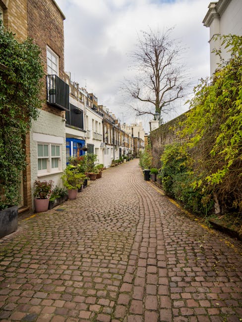 A narrow cobblestone street in South Kensington lined with a mix of residential buildings and greenery. On the left side, several potted plants and small trees are positioned along the base of the buildings, some with window boxes containing flowers. The buildings feature white and colorful facades with sash windows, and a black wrought iron balcony is visible on one of the upper floors. On the right side, a tall brick wall and a lush hedge with green leaves run parallel to the street, partially obscuring the buildings behind. At the end of the street, a large, leafless tree with bare branches extends upward, set against a cloudy sky. The street appears quiet and is likely used for local traffic or pedestrian access, fitting the context of a residential area suitable for house removals and home relocation services. Man with Van South Kensington would facilitate the loading process of furniture and boxes onto nearby delivery vans, utilizing the space within sight for efficient moving logistics, with some packaging materials like blankets or cardboard likely involved in protecting items during transport.
