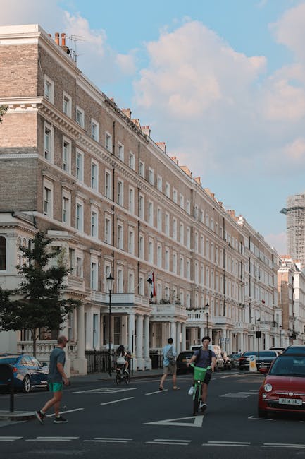 A row of elegant, multi-storey Victorian-style terraced houses with light-colored brick facades and white window frames along a busy city street under a partly cloudy sky. Ground-floor entrances feature columns and black railings, and some homes display small front gardens with trees and shrubs. In the foreground, several pedestrians are walking on the sidewalk, and a cyclist is riding along the street, while parked cars line the curb. The scene captures a typical urban environment where local residents and visitors are active, with a focus on residential architecture and street life. This image, associated with house removals and moving services, reflects the bustling atmosphere of South Kensington, relevant to regulations like council permits for moving vans, as promoted by Man with Van South Kensington.