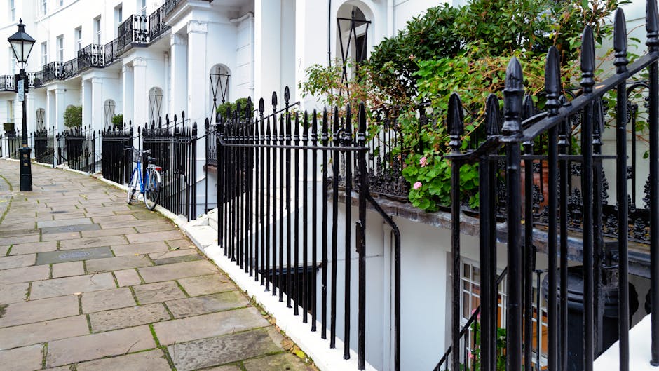 A image showing a residential street with white terraced houses featuring black wrought iron railings along the pavement. The sidewalk is paved with uneven stone slabs, and a blue bicycle is parked against the railings near a doorway leading to a lower basement entrance. Green foliage and pink flowering plants grow in pots and containers on the balconies and ledges of the houses. The scene appears to be outdoors during daylight, with natural lighting illuminating the scene. The image reflects a typical urban setting where house removals or moving services, such as those by Man with Van South Kensington, might occur, especially considering the need for parking permits and logistical planning for furniture transport and home relocation.