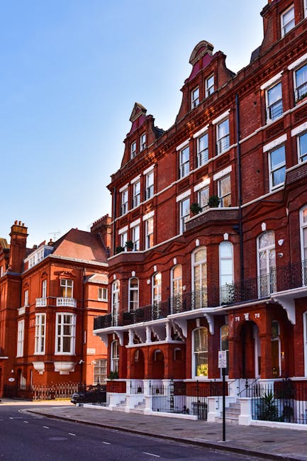 Photograph of a row of red brick Victorian-style townhouses on a city street, with multiple floors and black wrought iron balconies on the front. The buildings are tall with large sash windows, some with decorative stone lintels, and feature ornate architectural details near the roofline. The pavement in front of the buildings is clear, and there are no vehicles or people visible on the street. The sky above is bright and clear, indicating daytime. This residential area exemplifies classic London architecture, often involved in home relocation or furniture transport services. Man with Van South Kensington provides professional removals in such environments, where careful handling of furniture and boxes is essential during the loading process, typically carried out with equipment like trolleys and moving blankets to protect items during home moves.
