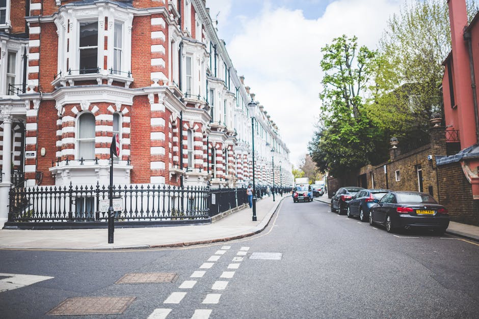 A street view in South Kensington featuring Victorian-style terraced houses with red-brick facades, white decorative window trims, and small front gardens enclosed by black wrought iron fences. Parked cars line the narrow, paved road, which has double yellow lines and white zigzag markings near the corner. A man with a moving box wrapped in plastic lies on a trolley, while another individual stands nearby, possibly engaged in home relocation or furniture transport activities associated with house removals. The scene is illuminated by natural daylight, with a tree providing greenery on the right side and a partly cloudy sky overhead. This setting aligns with the context of leaving or entering properties in an area where experts like Man with Van South Kensington handle packing, moving, and loading processes on narrow streets with limited parking access.