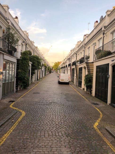 A narrow cobbled street in South Kensington with white 19th-century terraced houses on both sides, each featuring black railings and potted plants on balconies or window sills. A white van is parked in the middle of the street, positioned for home relocation and furniture transport. The pavement has yellow lines indicating parking restrictions, and a trolley or moving straps may be visible nearby as part of the loading process. The sky above is partly cloudy with soft evening light, suggesting the early stage of a house moving or packing and moving operation. This setting, characteristic of tight urban streets, highlights the logistical consideration for removals services, such as those provided by Man with Van South Kensington, in areas with limited parking access and narrow streets.
