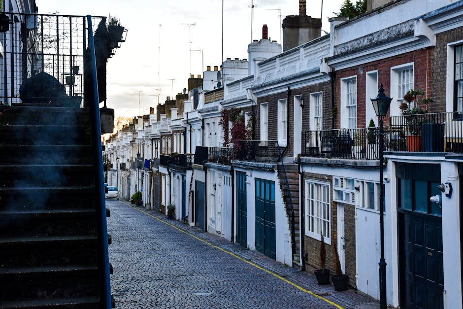 A street view in South Kensington featuring Victorian-style terraced houses with red-brick facades, white decorative window trims, and small front gardens enclosed by black wrought iron fences. Parked cars line the narrow, paved road, which has double yellow lines and white zigzag markings near the corner. A man with a moving box wrapped in plastic lies on a trolley, while another individual stands nearby, possibly engaged in home relocation or furniture transport activities associated with house removals. The scene is illuminated by natural daylight, with a tree providing greenery on the right side and a partly cloudy sky overhead. This setting aligns with the context of leaving or entering properties in an area where experts like Man with Van South Kensington handle packing, moving, and loading processes on narrow streets with limited parking access.