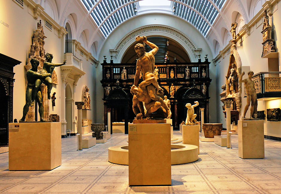 Interior view of a museum gallery displaying classical sculptures on beige pedestals arranged in a semi-circle, with a large central statue depicting a muscular male figure holding a smaller figure. The sculptures are made of bronze and marble, showing detailed human anatomy and drapery. The gallery features a high, arched ceiling with natural light streaming through skylights, white walls adorned with smaller sculptures and reliefs, and a wooden, ornately carved backdrop at the far end. Visible in the scene are various packing materials such as bubble wrap and plastic sheeting used for protection, as well as a black van parked outside the gallery door, indicating ongoing transportation or professional moving services. The environment suggests careful handling during a home relocation or museum transfer, with Manchester with Van South Kensington providing removals and logistics support for artwork and exhibit movements.