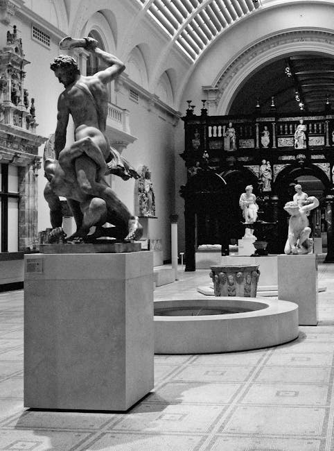 Black-and-white photograph of an interior space within the Victoria and Albert Museum, showcasing classical sculptures on display. In the foreground, a large marble sculpture depicts a male figure in a dynamic pose, partially draped, standing on a rectangular pedestal. Surrounding it are additional sculptures placed on similar pedestals, some featuring human figures in various poses. The room includes a decorative, ornate fireplace with intricate carvings on the left side, and a high, arched ceiling supported by a series of evenly spaced, curved beams. Light filters through the ceiling, illuminating the marble surfaces and the polished tiled floor. In the background, there is a dark wooden display case or partition with carved architectural details, housing smaller statues or artefacts. The museum setting is spacious and well-lit, with no visible visitors, and the focus remains on the classical sculptures, which are prepared for a home relocation or moving logistics process as managed by Man with Van South Kensington.